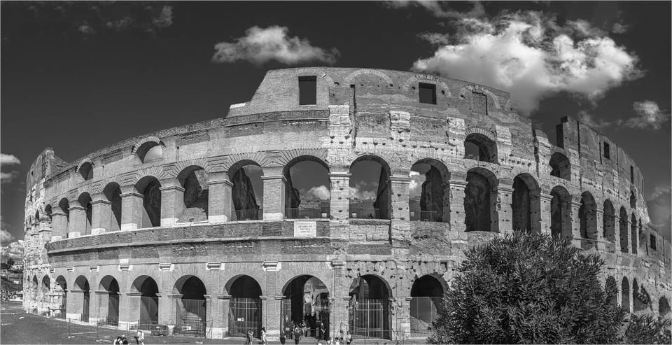 A black-and-white photo of the Roman Colosseum. It shows a large circular corroded ancient building, two stories, each story a series of arches. The stone is clearly extremely old but in quite good shape nonetheless