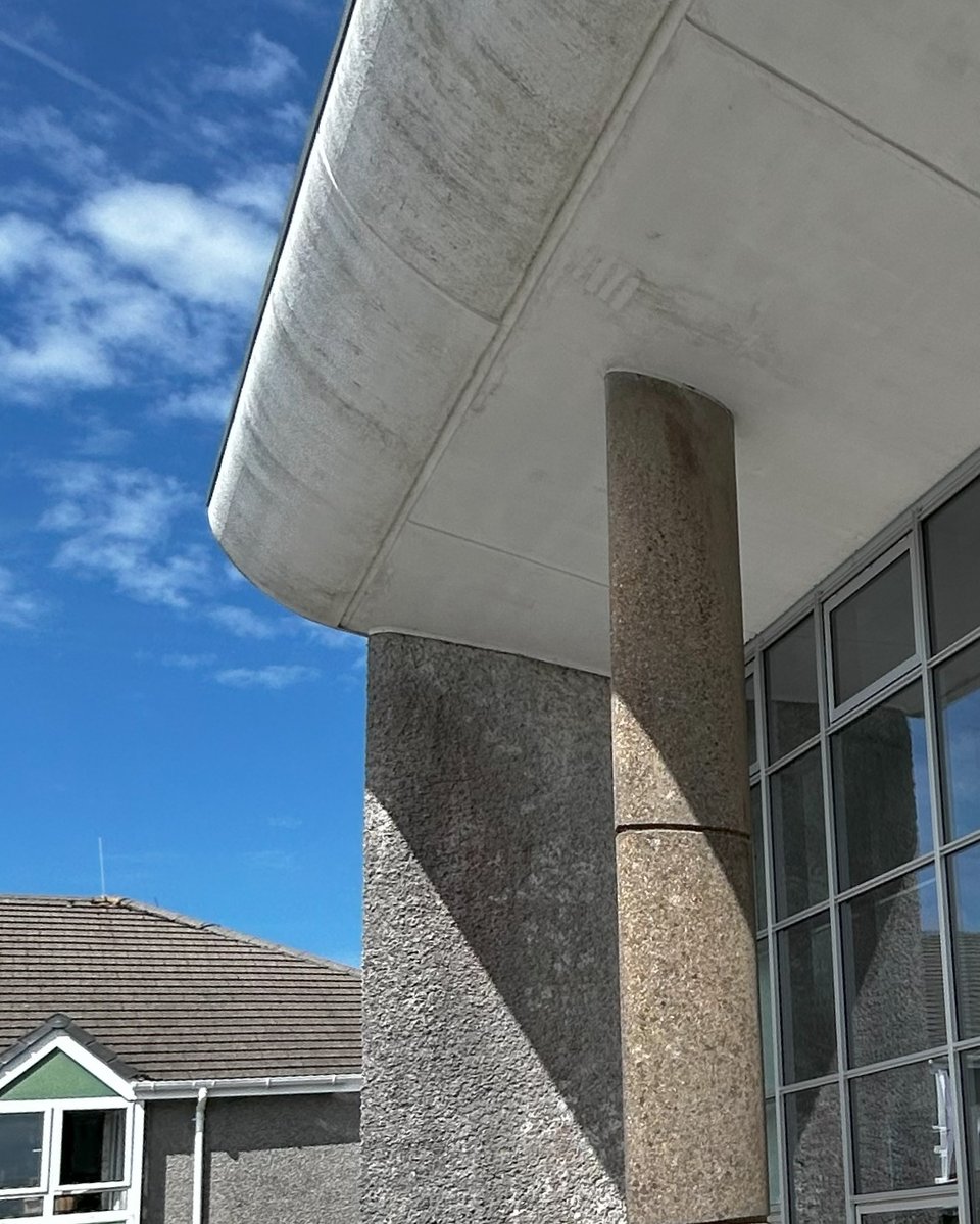 The white underside of a butterfly roof creating a deep porch. The supporting wall is of rough cast concrete and there a slender, oval pillar supporting the deep roof. A modern house is next to it all.