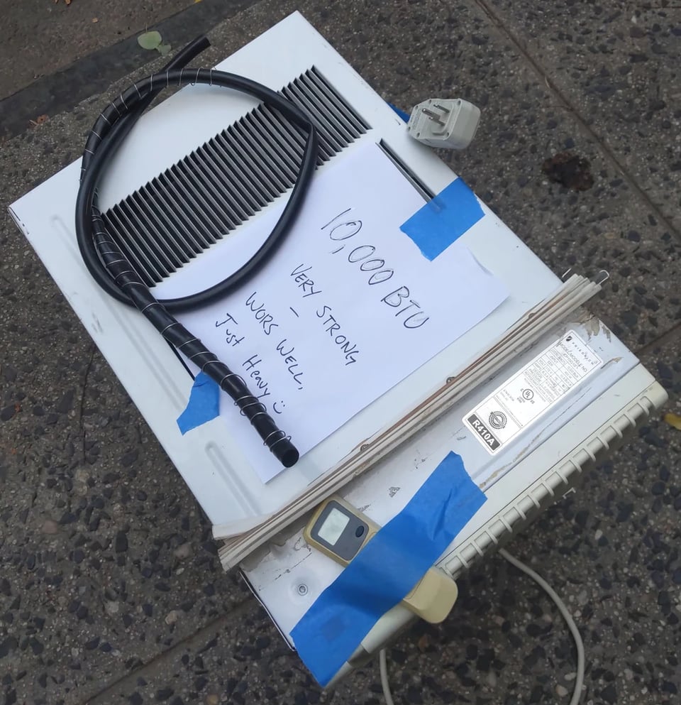 A large air conditioner on the sidewalk with its power cord and remote. There is a note taped to the top which says the AC has 10,000 BTU and works very well, but is just heavy with a smiley.