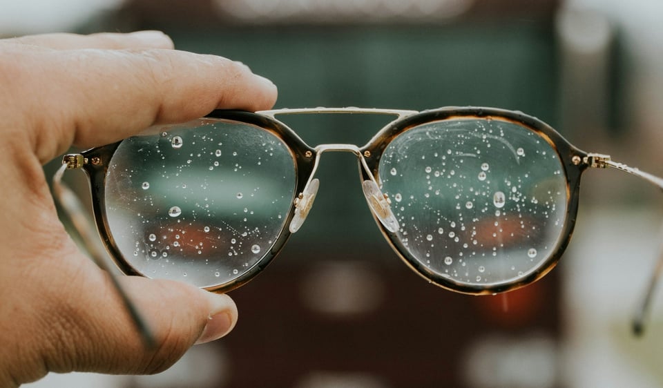 A hand holding up a pair of glasses. The lenses are streaked with droplets, making it difficult to see through them. Photo by Hermes Rivera on Unsplash.