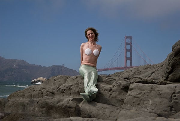 A white trans woman wearing a mermaid costume on some rocks with the Golden Gate Bridge in the background