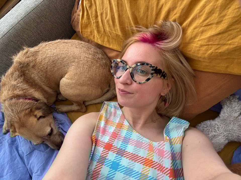A selfie with my dog, Momo. We are laying in bed together. I have a flash of magenta pink hair at my hairline and I look pretty comfortabled.