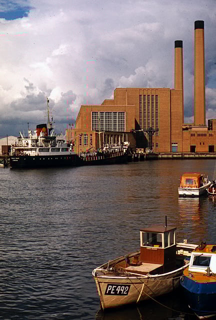A huge brick building with tall windows and two very tall chimneys sits by a seaside wharf. A coal cargo boat is offloading onto the wharf. In the foreground, a couple of small fishing boats are tied up.