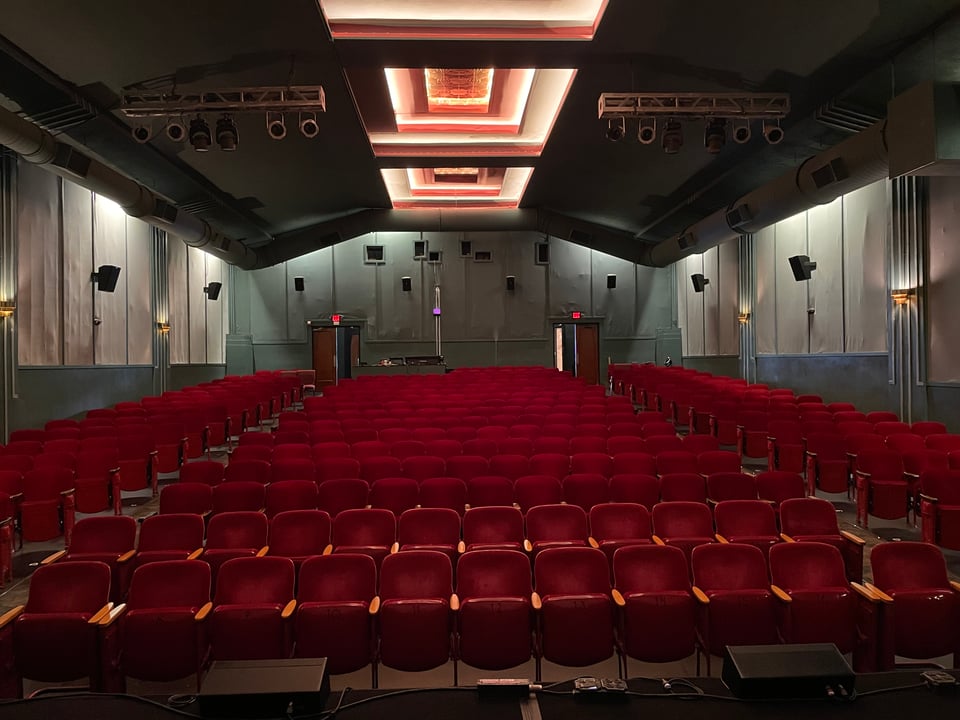 A theater viewed from the stage. A couple hundred red chairs and art deco lights on the ceiling