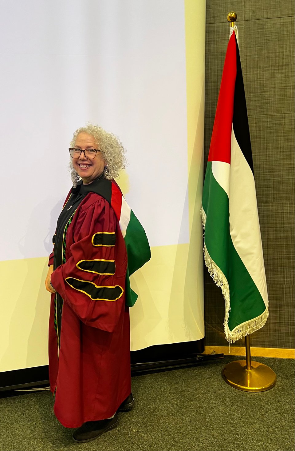 A woman in red academic regalia, next to a Palestinian flag. The hood of the regalia is in the colors of the flag