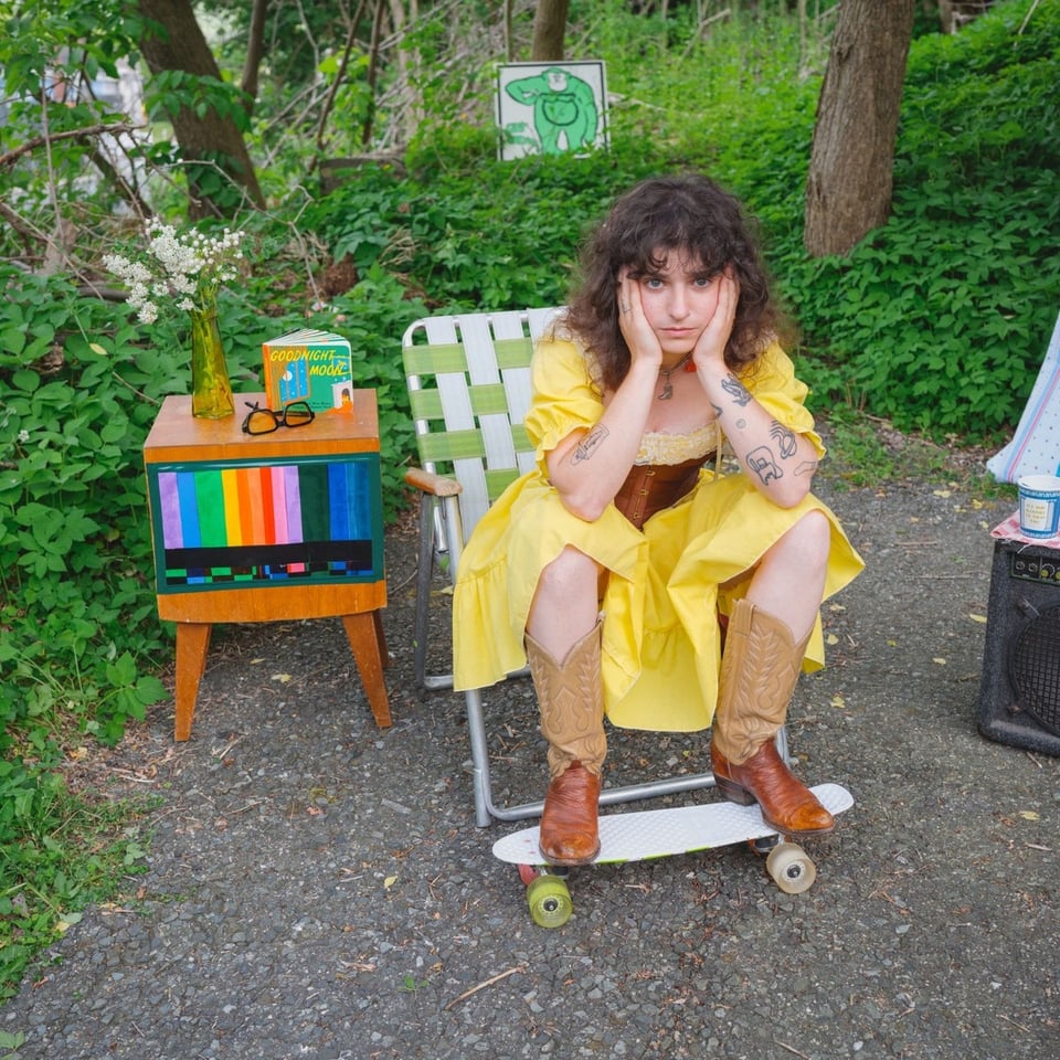 W woman in a yellow dress and cowboy boats sits in a folding lawn chair with her hands on her cheeks and her feet resting on a skateboard.