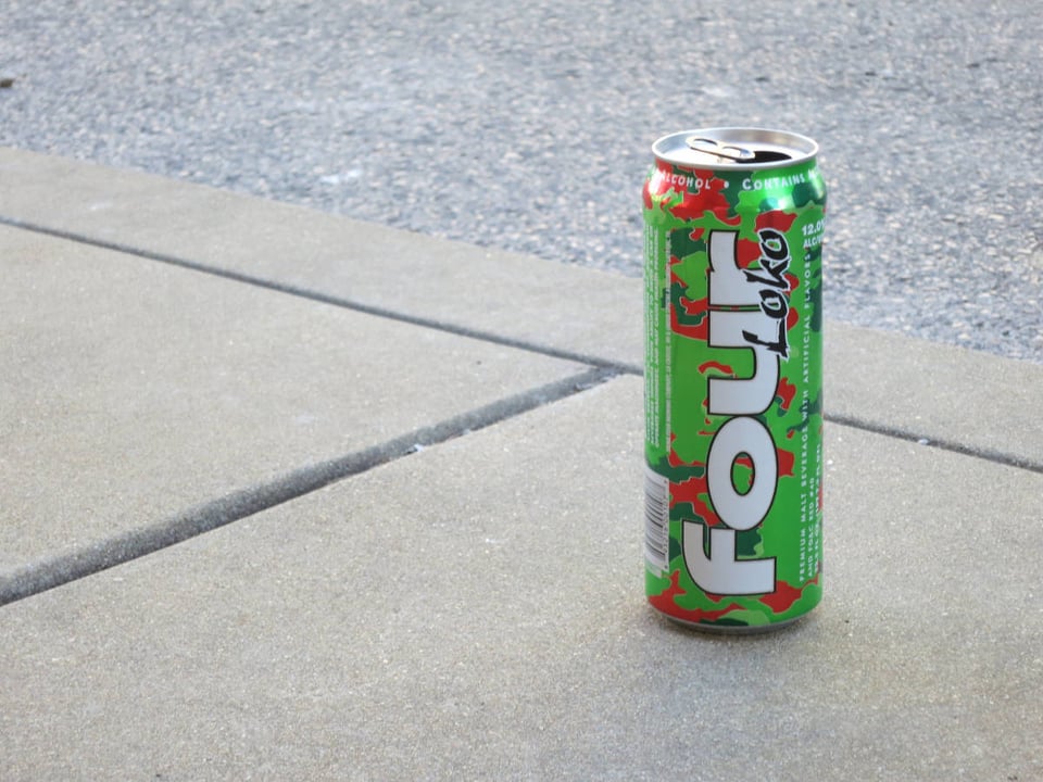 A photo of a can of Four Loko. It is tall and green, with its logo name in white, and it is sitting on a sidewalk with a bit of road visible behind it.