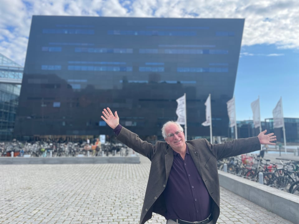 A man in a jacket raises his arm in happiness as he stands in front of the "black diamond" Public Library in downtown Copenhagen.