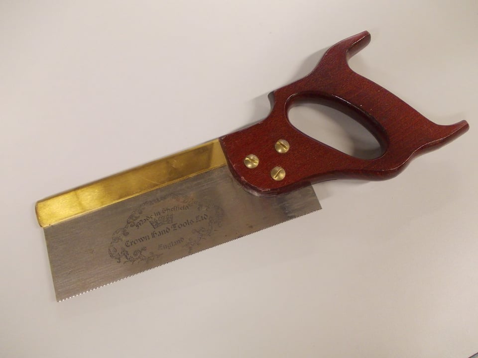 A hand saw about 12" long with many small teeth, a bras metal fitting on the top of the blade, and a red/brown handle.