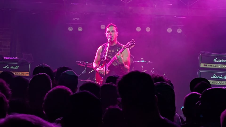 A shot from the crowd of a white man, Chris Hannah of the band Propagandhi, playing a red Gibson SG guitar, surrounded by amps, standing in front of the drummer, bathed in purple light.