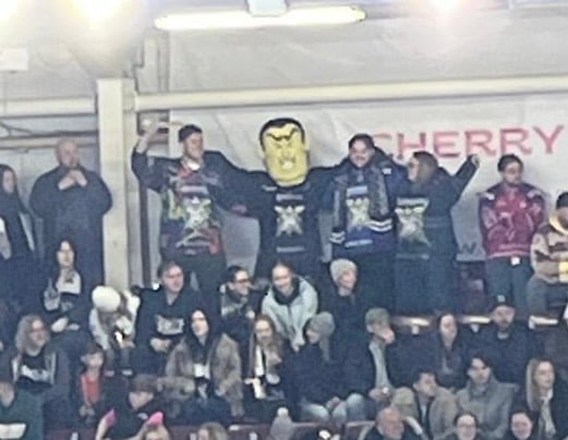 The above man (bit yellow foam head) standing in the back row of tiered seating, with his arms on the shoulders of the men standing either side of him.