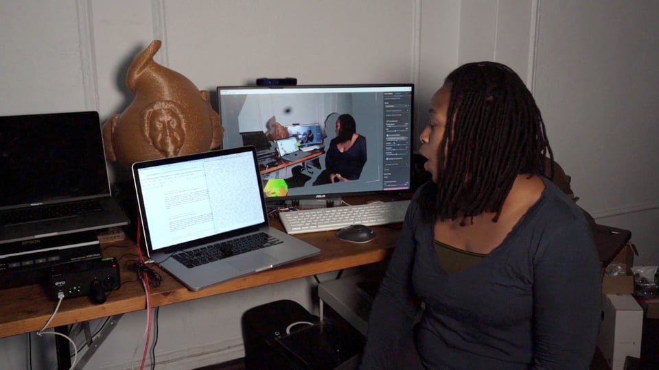 Stephanie at her work station. She speaks toward a laptop with a text window open, and behind it is a bigger monitor showing a video stream of Stephanie speaking into the laptop. Also behind the laptop is what looks like a large golden gourd or cornucopia with sculptural faces emerging from each side. That’s N’TOO’s physical manifestation.