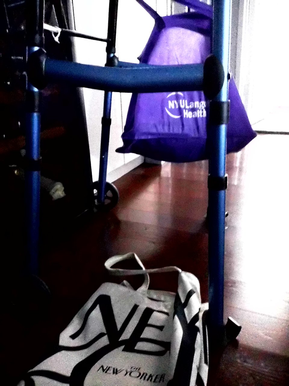 a digital Instax photo of a blue metal walker, with a purple NYU Langone Health bag hanging from the handle, and a New Yorker tote bag on the floor