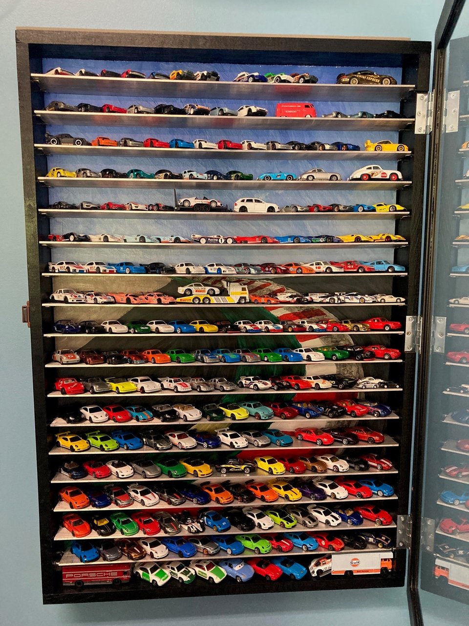 A display case with aluminum shelves containing numerous diecast vehicles. The background is a painting.