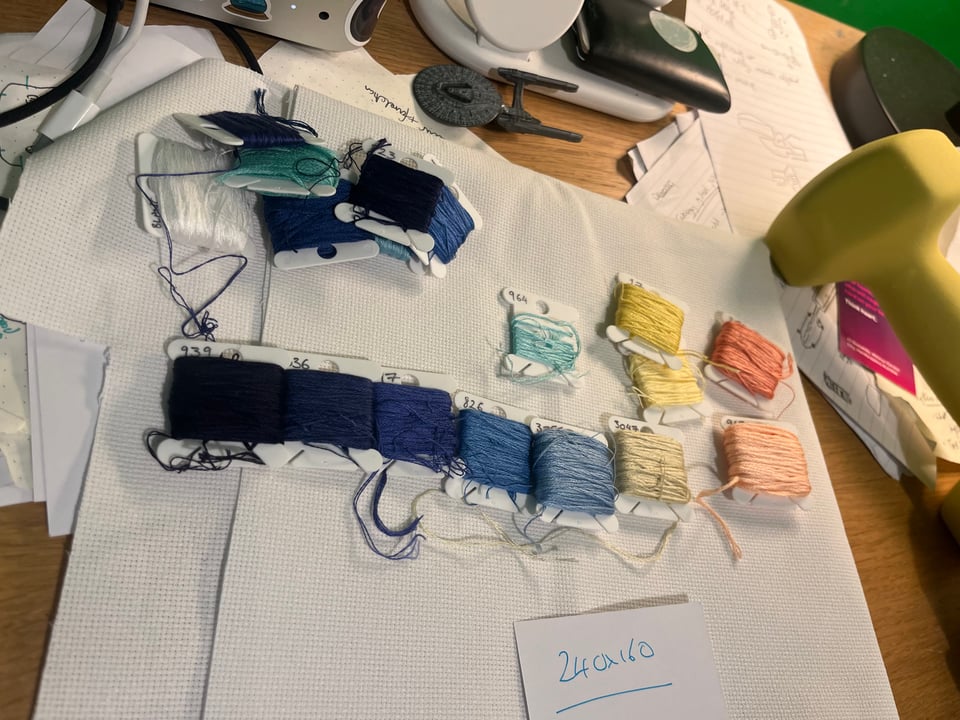 Some cross-stitch fabric covered in coloured bobbins arranged into a gradient of blue and orange, and other desk mess around them.