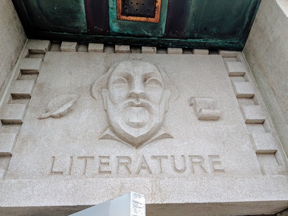 It's a decoration on the wall of a high school. There is  a man who may be William Shakespeare, hard to say, with a mustache and a pointy beard. He looks like his mouth is open very wide but I think that's just the mustache and beard creating an optical illusion. There's a leaf on his left and a scroll on his right. Underneath it says LITERATURE.