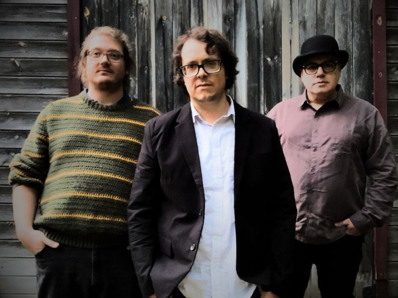 Three men comprising the band Ginger Bug, each wearing glasses, stand together against a backdrop of weathered wood.