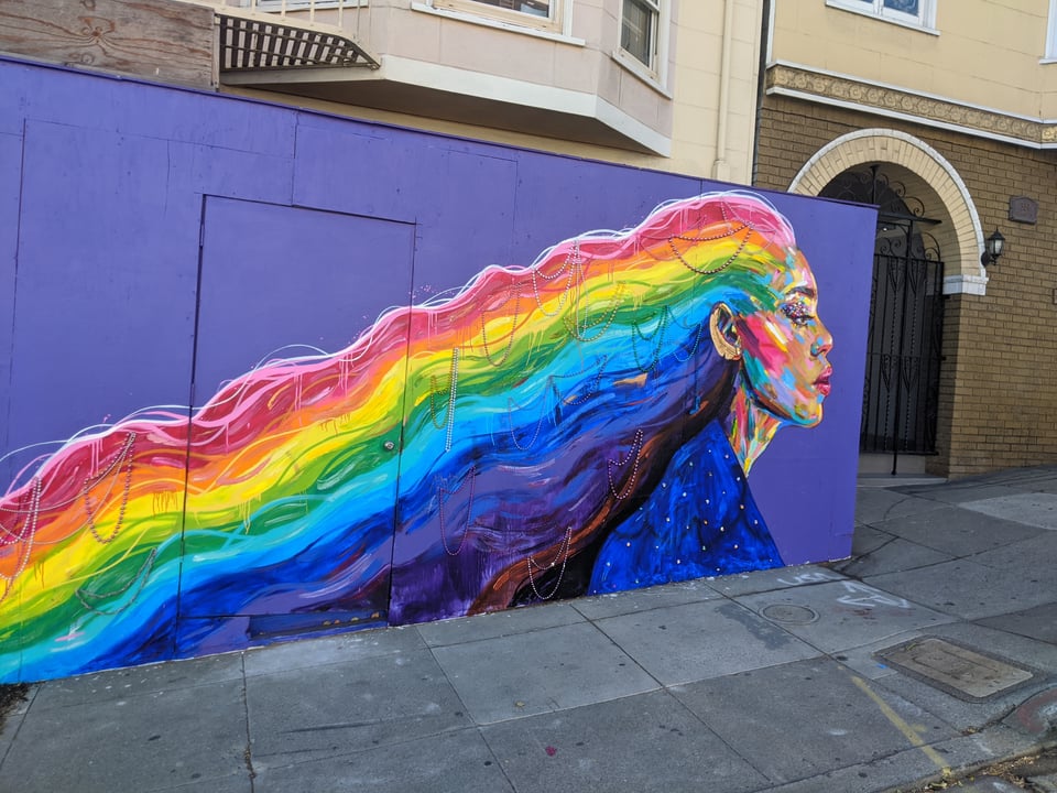 A mural of Laverne Cox, a Black trans woman, with rainbow hair streaming behind her, wearing a dress that looks like the night sky. She' sagainst a purple background.