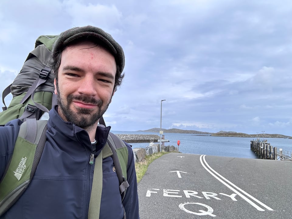I stare at the camera wearing a big green backpack that peaks over the top of my cap. Behind me is a road leading to the docking area for a ferry. The ferry has not yet arrived. Mountains and islands are visible across the water.