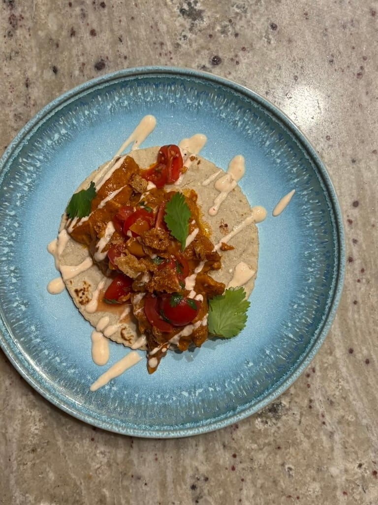 A white corn tortilla drizzled with a yogurt-chipotle sauce and topped with peanut-stew chicken, a tomato-scotch bonnet salad, cilantro, and crispy bits of chicken skin on a blue plate.