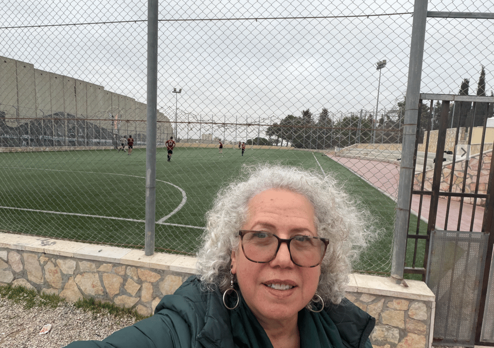 Photograph of a woman with curly gray hair and glasses taking a selfie in front of a fenced soccer field with players in the background, under a gray sky. Next to the field is a large concrete wall.