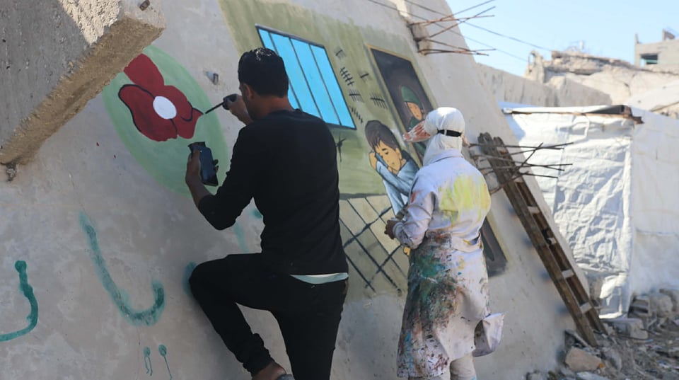 Two artists photographed from behind as they paint a mural on a wall, one illustrating the Heal Minds Gaza logo, and the other painting the mural of the child crouching in a prison cell. In the background are tent tarps and buildings