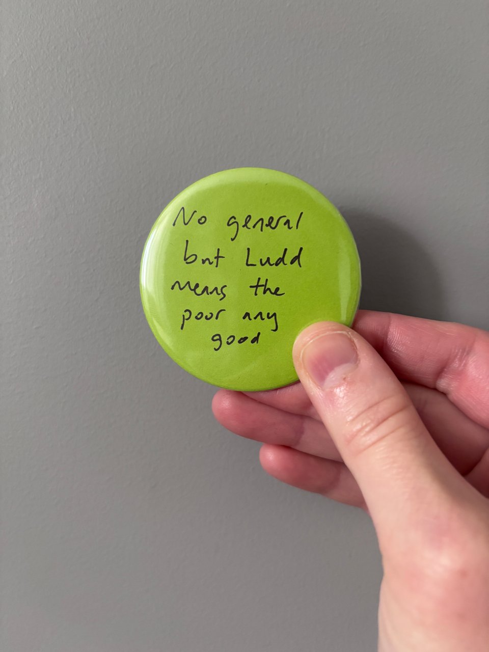 a light-skinned hand holds a lime green button pin that says "No general but Ludd means the poor any good" in black ink