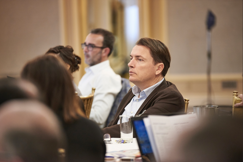 Photo from a business conference with nearby figures blurred and focus on a man in shirt and jacket, Jeroen van der Linden, listening intently to a conference speaker.