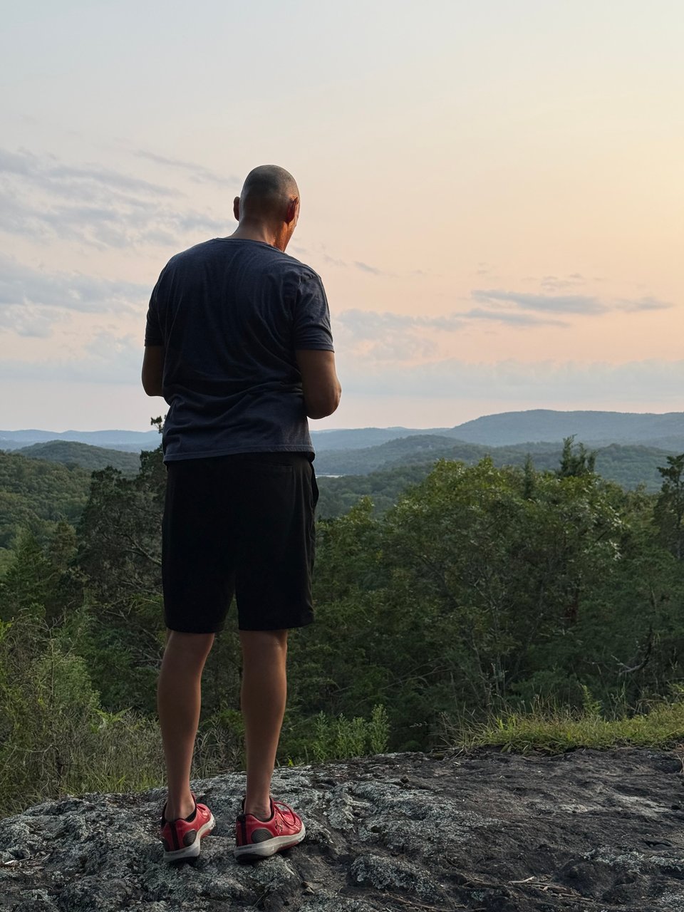 photo of a tall white man with a shaved head, standing atop a rock cliff, with trees and hills extending far into the background. He is in a blue T-shirt, black shorts, and red sneakers, and is looking down at his phone. He's somewhat silhouetted by the sunset light off to the right of the frame