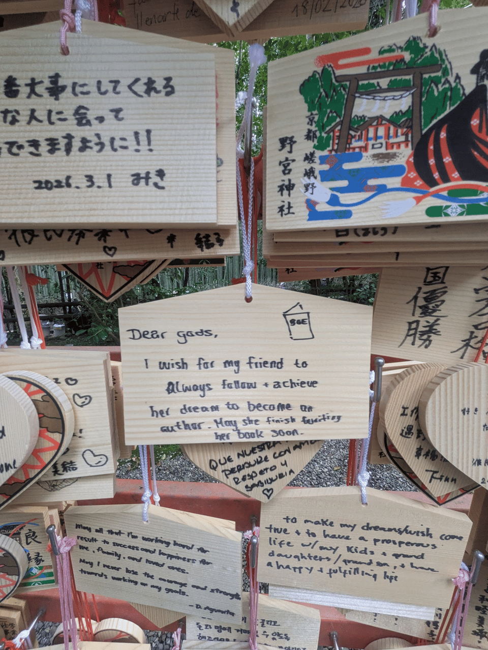 A group of wooden emas with black sharpie-written wishes. There are many in the picture including Japanese, English, Spanish, Korean, and Mandarin. The central one reads, "Dear gods, I wish for my friend to always follow + achieve her dream to become an author. May she finish writing her book soon." It also has a small picture of a book with the letters "BGE" on the cover.