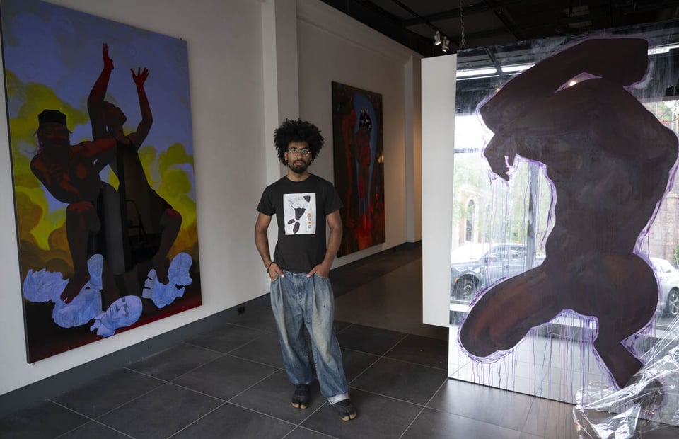 A man with an Afro and eyeglasses wearing a black t-shirts and blue jeans stands in a gallery flanked by artwork.