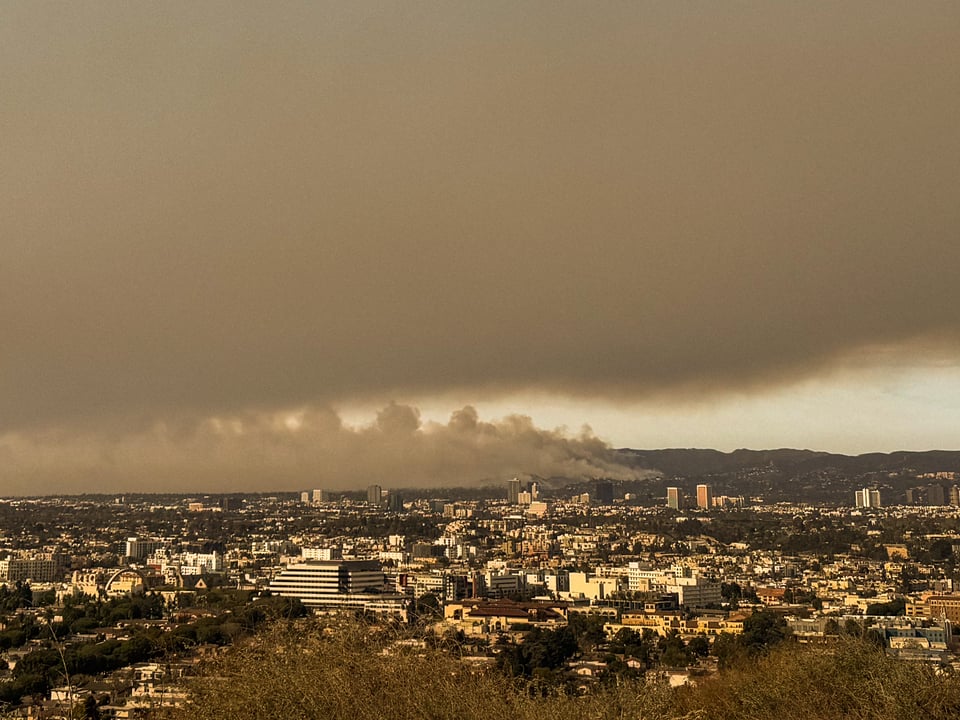 A hilltop view of Los Angeles with the Santa Monica mountains visible beyond. Smoke rises from those mountains, and a large, dense cloud of smoke floats above