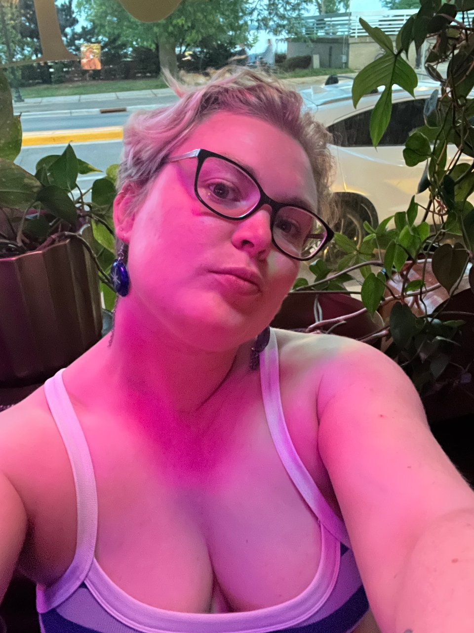 Nola pursing her lips at the camera, hair swooped back and to one side, wearing dangling bottle-top earrings and a low cut striped stretch tank, plants and a window in the background