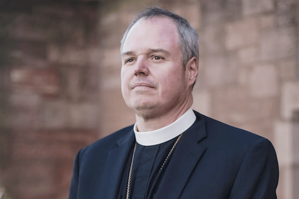 a white early middle aged man with in black shirt and jacket with Anglican clerical collar