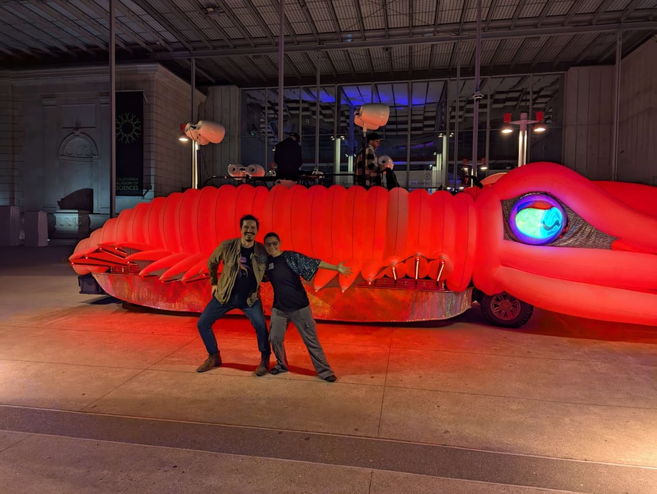 A photo of me (Danna Staaf) and Sam Julian posing in front of the Sepia Lux art car. At the moment of the photo, this giant cuttlefish-shaped vehicle is glowing red, with the W-shaped pupil of a cuttlefish visible on the screen that makes up its eye. Sam and I look justifiably excited.