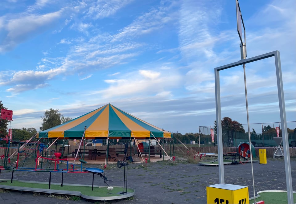 A scene from a mini golf arena, with yellow and green round top circus like tent (without the side walls) sitting area being prominent on the left side. There are different mini gold contraptions visible with mechanical pathway and wind controlled tunnels scatterred around. The scene is from a late afternoon