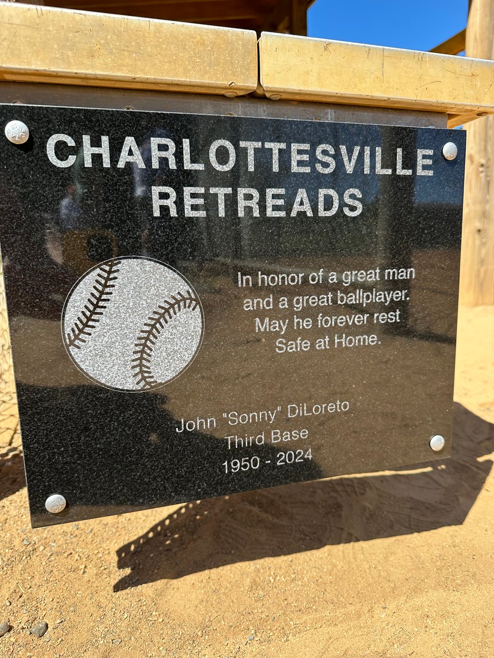 Photo of a plaque at the end of the home team's bench on a softball field. It reads: