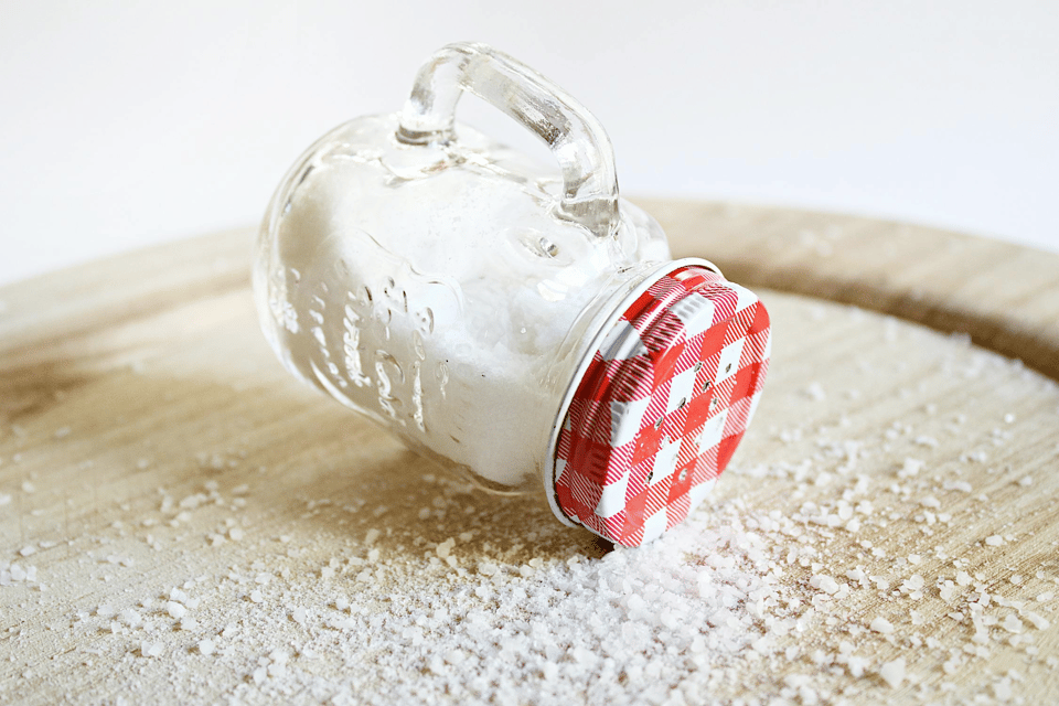 A photo of a salt shaker fashioned from a small glass jar that has a handle and a metal lid with holes cut into it. The lid has a red and white gigham pattern. It is lying sideways on a wooden table and coarse salt is scattered all around.