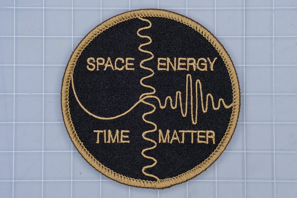 Circular embroidered patch with the quadrants labeled: space, energy, time, matter, inside a modified yin-yang symbol with waveform patterns