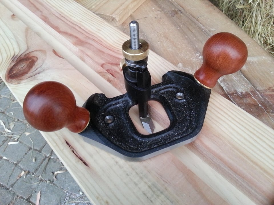 A router plane - a piece of metal with two round wooden knobs on either side and a blade in the middle. The blade is pointed and beveled and sits below the plane of the metal base. The plane itself is on top of a piece of wood that has a groove cut into it where the blade sits.