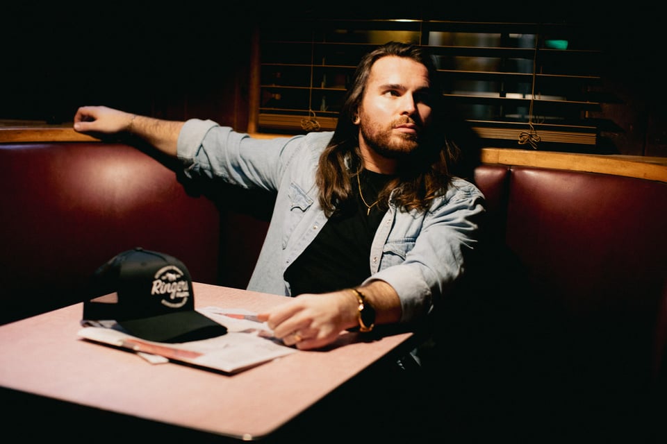 A man with a beard and hair past his shoulders sits at a banquette with a trucker cap on the small square table.