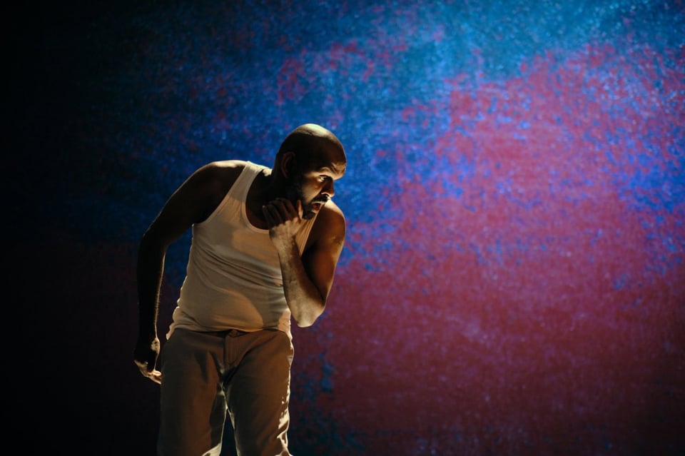 Ramesh Meyyappan, wearing a white singlet and white slacks, bends slightly and cradles his chin against a blue and red paint-splattered background.