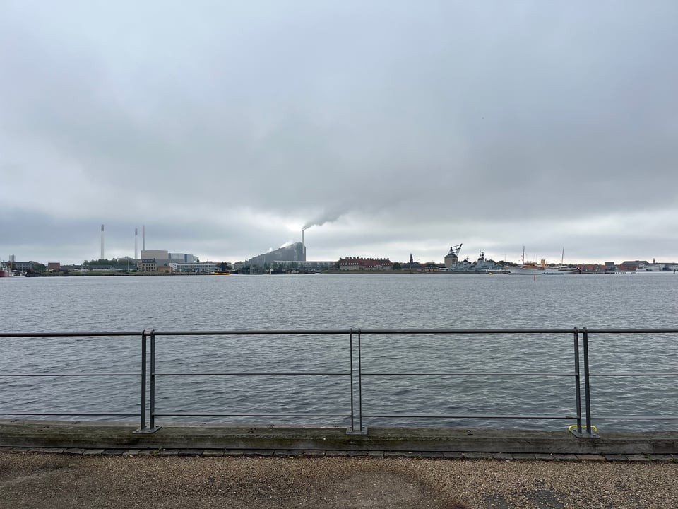 Past a fence lies a wide expanse of gray water and beyond it a skyline with what looks like a factory, with a smoke stack. Copenhagen Harbor.