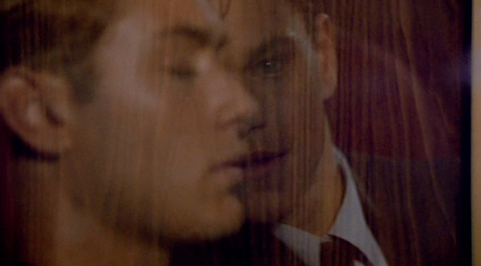 Two men's faces reflected in a window, one in profile and the other behind the first and facing forward.