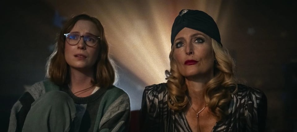Two women sit side by side gazing upward with awestruck expressions as bright rays of light radiate from behind them — one wearing glasses and a chunky green knit sweater, the other in a black turban-style headwrap and patterned dark blouse with blonde curls framing her face.
