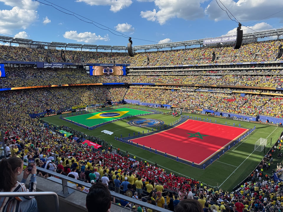 Photo of Brazil & Morocco’s flags laid out on the pitch at MetLife stadium before their match; the stands are filled with 80,000 fans, many in yellow Brazil kit