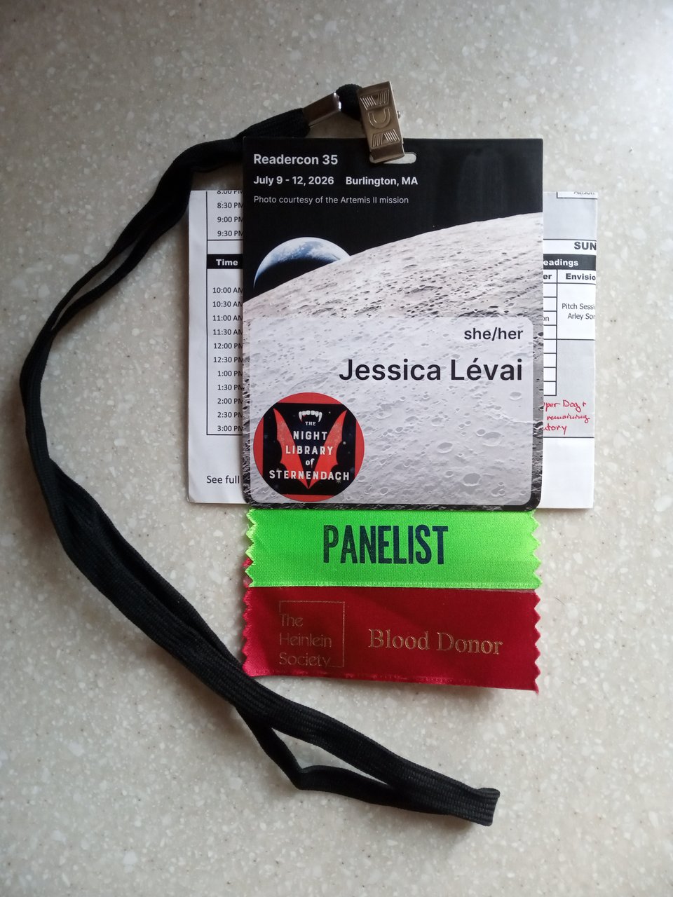My ReaderCon 2026 badge. The badge has my name and pronouns on a background of the lunar surface, with a sticker of the Sternendach cover affixed below. There is a green ribbon that says “Panelist” and a red ribbon that says “Blood Donor"