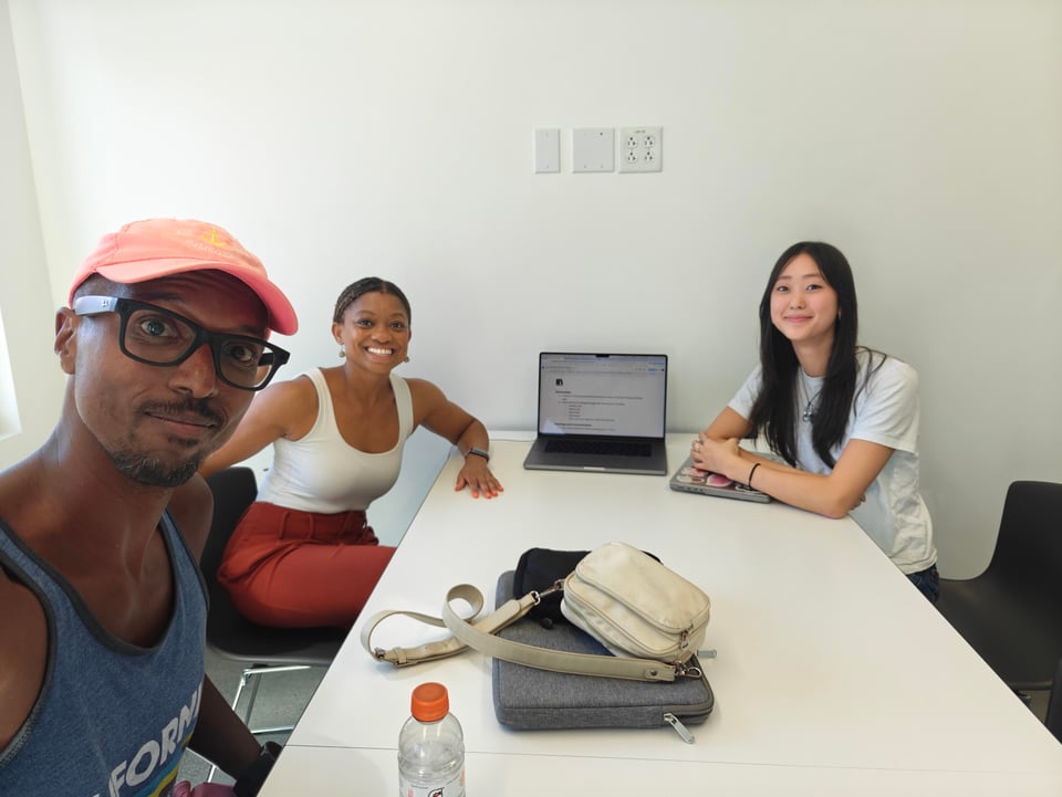 Rob, Nadia, and Yeji sit around a white table, smiling into the camera. At the far end is an open laptop.