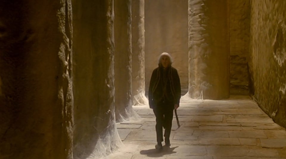 A man with long grey hair and a black suit circa the 17 or 1800s, in a stone hall way with rough pillars, cobwebs at their bases
