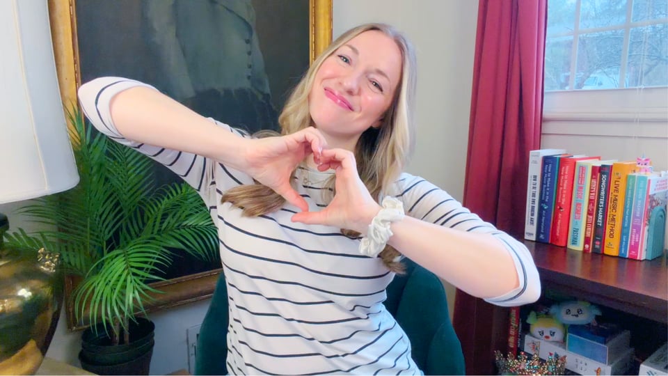 A picture of Emily Henry smiling and making a lopsided hand heart.